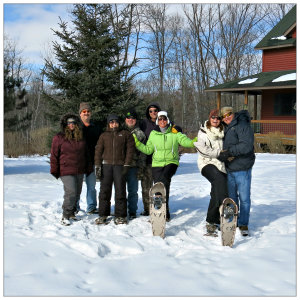 A group of people standing in the snow on snowshoes with a brown cabin in the background