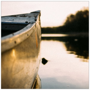 Front tip of a grey canoe on calm water at sunset with trees in the background 